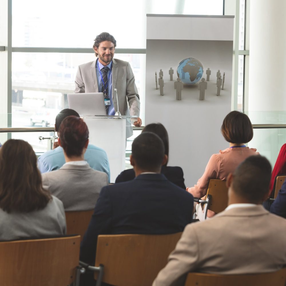 Front view of Caucasian businessman with laptop speaks in front of diverse group of business people sitting at business seminar in office building Front view of Caucasian businessman with laptop speaks in front of diverse group of business people sitting at business seminar in office building
